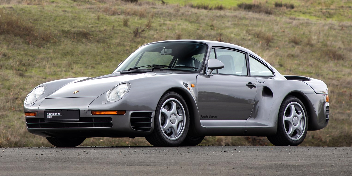 A grey Porsche 959 parked in front of a hill