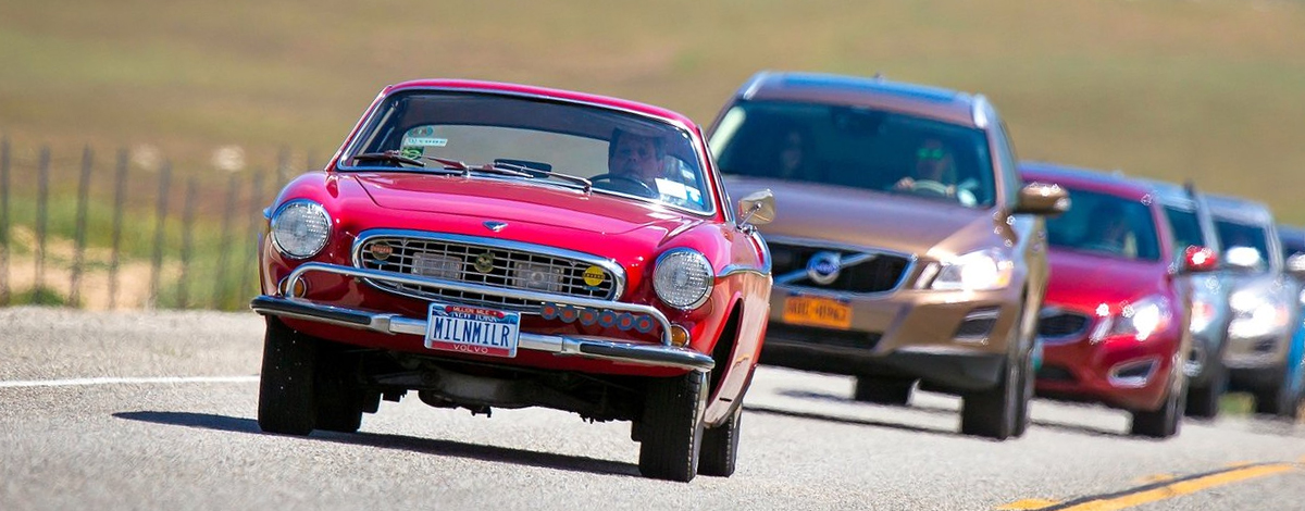 A red Volvo P1800 classic car driving in front of a line of traffic behind
